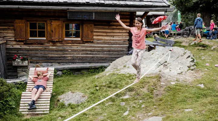 Zwei Kinder balancieren auf einem Seil, das vor einer Berghütte gespannt ist | © DAV/Hans Herbig